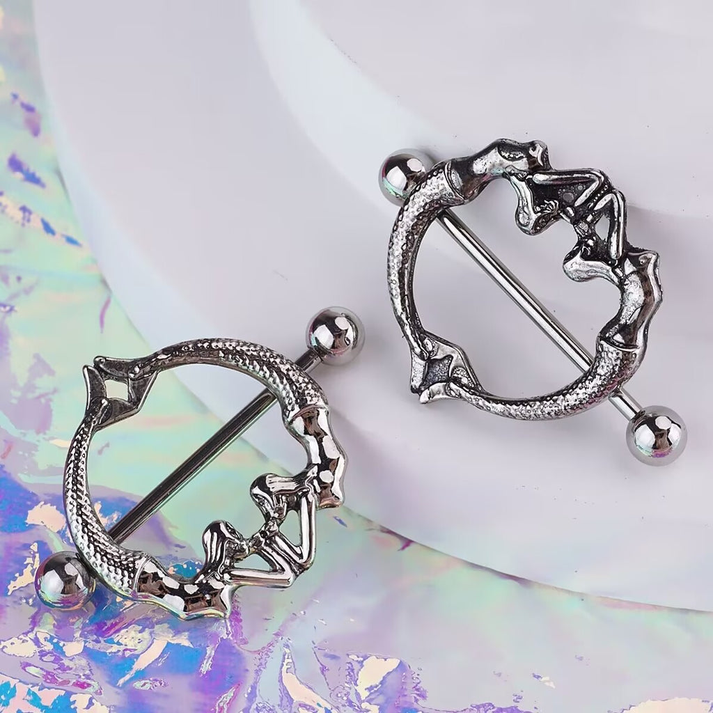 Two silver gothic nipple shields with anatomical pelvis design on a holographic background.