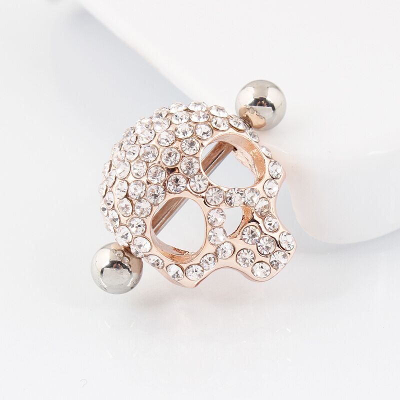 A profile view of a rose-gold skull nipple shield resting on a white surface, showing the barbell and side profile.