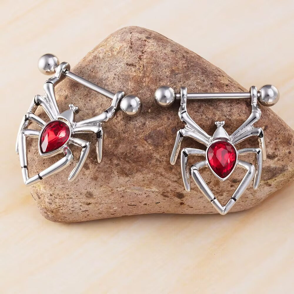 Pair of polished silver spider nipple piercing shields with red gemstones resting on a natural stone.
